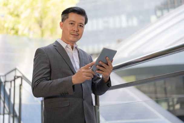 Уверенный бизнесмен в умном костюме занимается своим планшетным компьютером, стоя на открытом воздухе в современной городской обстановке, представляя связь и мобильные технологии в бизнесе - Фото, изображение