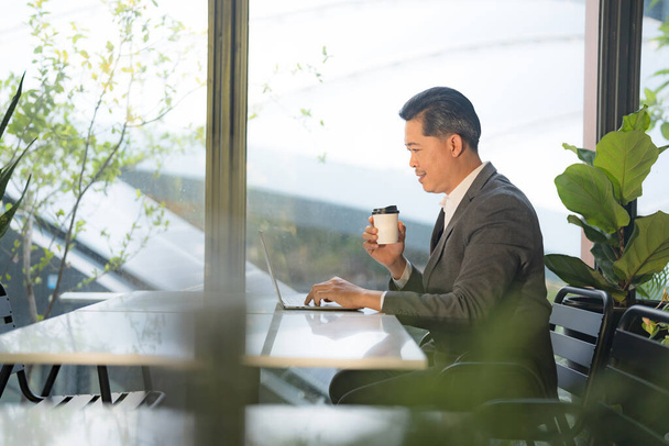 Gericht mannelijk bedrijf professionele werken op zijn laptop in een outdoor cafe, omgeven door groen en natuurlijk licht, een voorbeeld van mobiel werk-leven evenwicht in een stedelijke omgeving - Foto, afbeelding