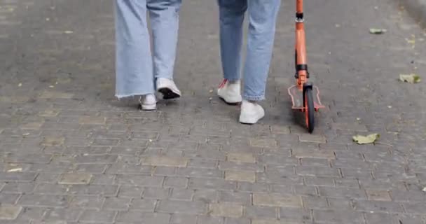 Nastolatki w dżinsach chodzą po drogach brukując kamienie hulajnogą. Sportowe dziewczyny spacerują w centrum miasta mówiąc o przyszłych planach sportów ekstremalnych - Materiał filmowy, wideo