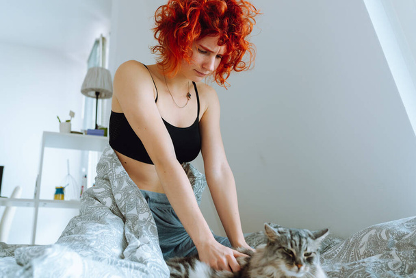 смішна руда дівчина-підліток, вранці в ліжку, масажує домашню кішку, обіймає і цілує кота Мен Куна. Дружба між домашнім улюбленцем і його власником - Фото, зображення