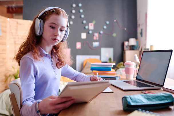 Mediana toma de la chica adolescente caucásica moderna con auriculares sentados en el escritorio estudiando en casa en su dormitorio, espacio de copia - Foto, Imagen