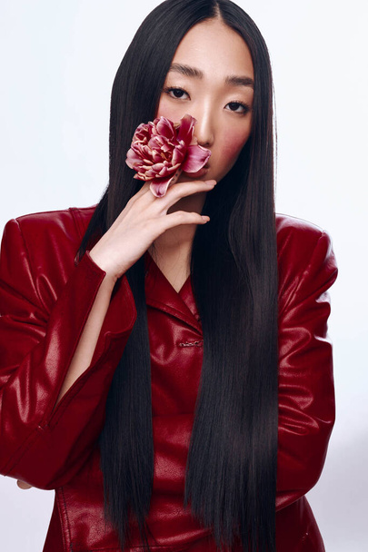 ピンクの花を保持する赤い革ジャケットの長い髪のスタイリッシュなアジアの女性 - 写真・画像