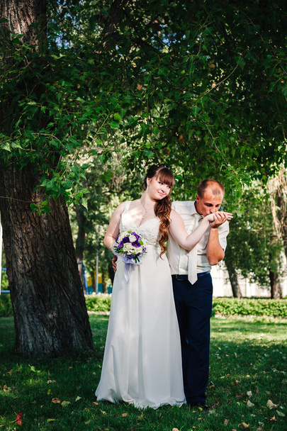 wedding couple kissing in green summer park. bride and groom kissing, standing together outdoors, hugging among green trees. Bride holding wedding bouquet of flowers - Photo, image
