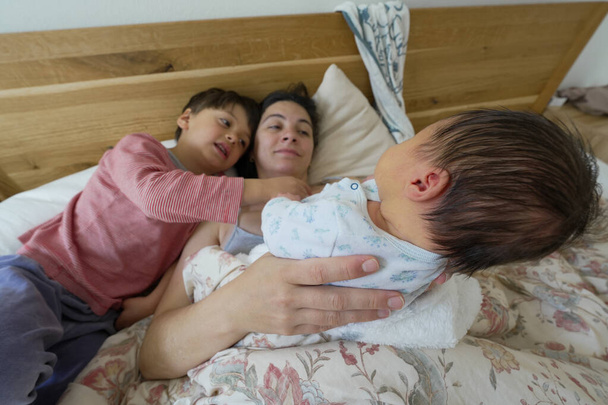 Madre acostada en la cama con su bebé recién nacido y su hijo mayor, interactuando amorosamente, destacando el vínculo familiar y el cuidado gentil compartido entre ellos en un ambiente cálido y hogareño - Foto, Imagen