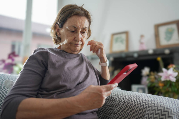 Seniorin mit kurzen Haaren trägt graues Hemd, sitzt auf einer Couch und betrachtet ihr rotes Smartphone mit nachdenklichem Blick, Inneneinrichtung mit heller Beleuchtung und Wohnkultur - Foto, Bild