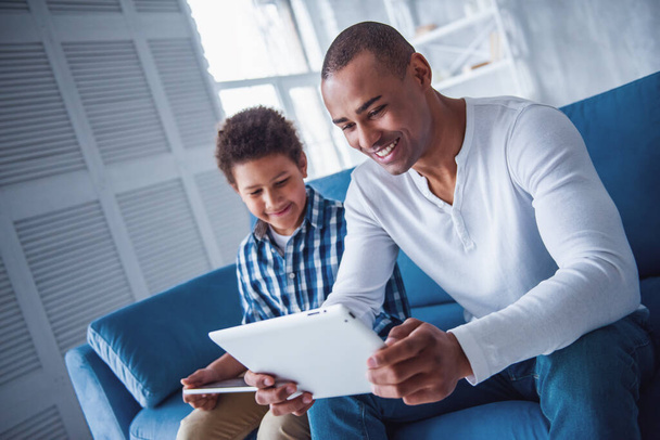 カジュアルな服を着た幸せな父親と息子は,自宅でソファーに座っている間にデジタルタブレットと微笑みを使用しています - 写真・画像