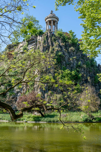 Paris, France - 05 11 2024: Park Buttes-Chaumont. View of The Sophora of Japan tree, the belvedere island, Temple of the Sibyl and the lake behind - Photo, Image
