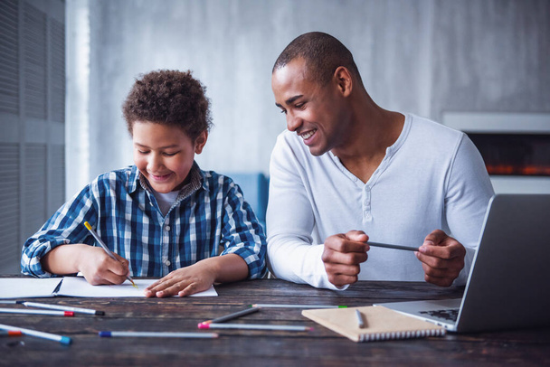 カジュアルな服を着た幸せな父親と息子は,自宅でテーブルに座っている間,描き,微笑んでいる - 写真・画像