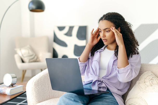 Exhausted brazilian or hispanic curly haired woman, freelancer or interior designer, sitting on comfortable sofa with laptop, sadly looks at screen, worried about work result, stressed about deadline - Fotografie, Obrázek