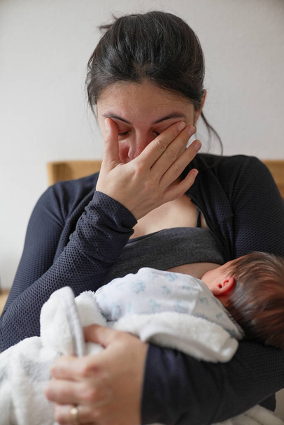 Madre esausta che allatta il neonato, asciuga le lacrime, rappresenta le sfide e le emozioni della maternità precoce. Questo momento grezzo ritrae la lotta e l'amore nel periodo post-partum - Foto, immagini