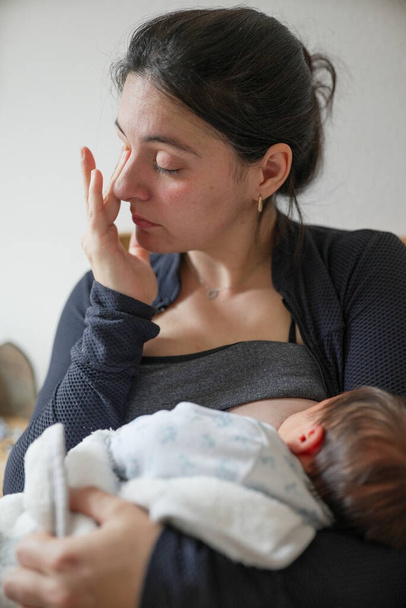 Új anya, megtörli a szemét, miközben szoptatja a gyermekét, megragadja a korai anyaság érzelmi és fizikai kimerültségét. Az új szülők előtt álló szeretet és kihívások szívből jövő ábrázolása - Fotó, kép