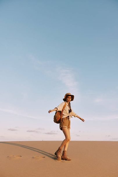 Жінка в капелюсі і шортах досліджує величезний пустельний пейзаж, прогулюючись через піщану дюну під палючим сонцем - Фото, зображення