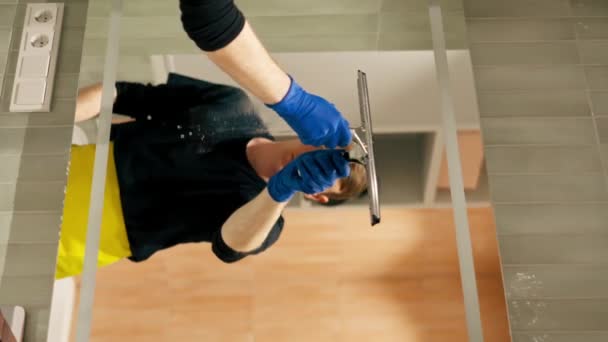 вертикальное видео в квартире уборщик в желтом фартуке чистит зеркало резиновой щеткой - Кадры, видео