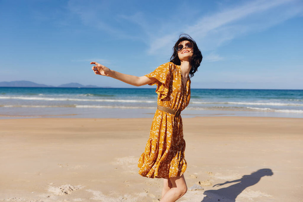 Женщина в жёлтом платье с вытянутыми руками, стоящая на пляже с безмятежным выражением лица на фоне прекрасного океана - Фото, изображение