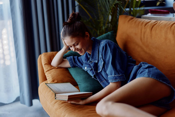 Young woman relaxing on couch with legs crossed reading an open book in cozy home environment - Photo, Image