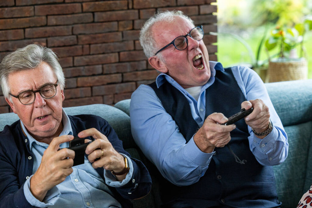 Відвертий постріл двох літніх чоловіків інтенсивно займався грою в відеоігри, сидячи на дивані. Один чоловік, одягнений в окуляри і синю сорочку, зосереджений на контролері, а інший - Фото, зображення