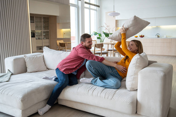 遊び心のある楽しい夫は敏感な喜びの妻をくすぐる. 現代のアパートのソファーで枕で男と戦っている笑う女性. 楽しい,喜び,幸せ,ポジティブな感情を持つ幸せな愛する家族 - 写真・画像