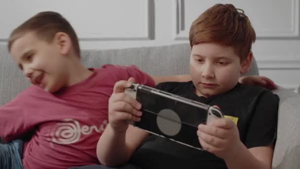 Un pequeño hermano menor mirando cómo su hermano tocaba la consola de mano. Dos chicos disfrutando de videojuegos juntos en el salón. Típico gasto de tiempo libre de los niños modernos. Saliendo de la habitación, cuando hermano - Metraje, vídeo