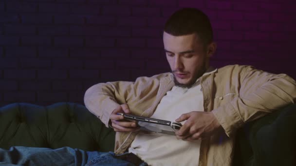 屋内でポータブルコンソールでビデオゲームをしている若い男. 男性はタブレットを使用してモバイルゲームを楽しんでいます. 自宅でゲームでひとりで過ごす時間. 若い男はポータブルコンソールでゲームを選択するリラックス - 映像、動画