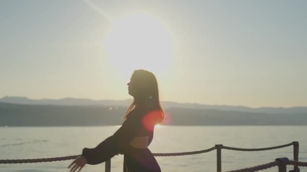 La femme est heureuse debout sur la jetée, regardant le coucher du soleil sur l'océan. Le ciel est rempli de lumière du soleil chaude, créant un bel horizon au crépuscule. 4K Vidéo de haute qualité - Séquence, vidéo