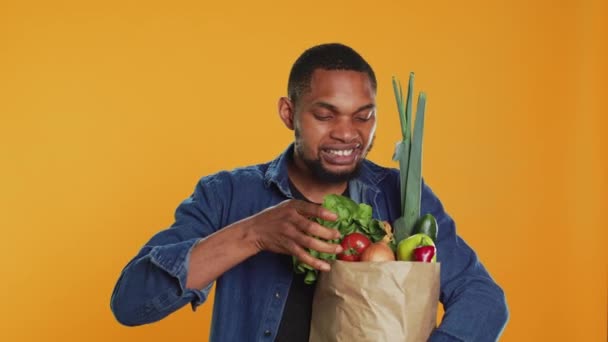 Onnellinen nuori mies mainostaa luomu paikallisesti kasvanut hedelmiä ja vihanneksia, esittelee biokemikaaleja ilmainen porkkana studiossa. Tyytyväinen ihminen tukee terveellistä ruokavaliota ja vegaanisuutta. A-kamera. - Materiaali, video