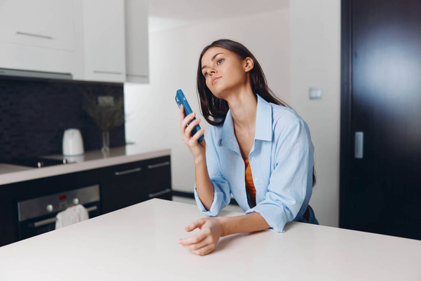 Frau sitzt mit Handy in der Hand am Küchentisch und blickt in lässiger häuslicher Umgebung in die Kamera - Foto, Bild