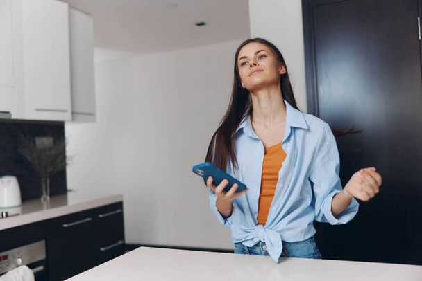 Кавказька жінка стоїть на кухонній стійці з руками в повітрі, тримаючи мобільний телефон - Фото, зображення