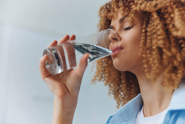 Mulher com cabelo encaracolado Desfrutando de uma bebida refrescante de água em um copo em uma sala brilhantemente iluminada - Foto, Imagem