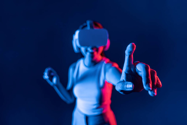 Inteligentna kobieta stojąca w cyberpunkowym neonowym świetle nosi zestaw słuchawkowy VR łączący metawertyczną, futurystyczną technologię społeczności cyberprzestrzeni. Elegancka kobieta używa wirtualnego obiektu wygenerowanego przez palec. omamy. - Zdjęcie, obraz