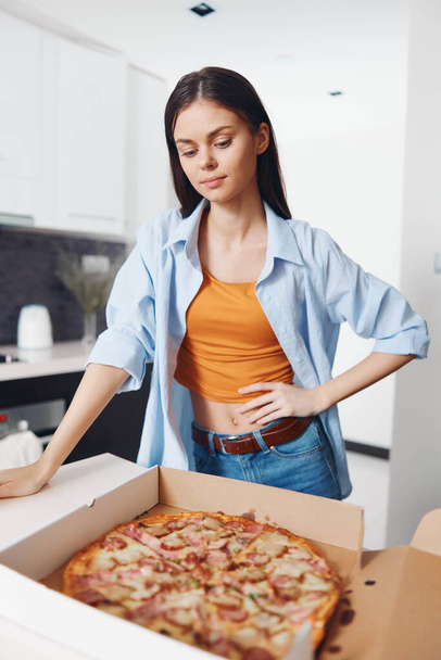 Γυναίκα στέκεται μπροστά από ανοιχτό κουτί πίτσα με πεπερόνι και τυρί toppings σε αυτό στη ρύθμιση της κουζίνας - Φωτογραφία, εικόνα