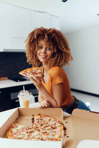 gelukkig jong Afro-Amerikaans vrouw met krullend haar genieten van een plak pizza in een doos - Foto, afbeelding