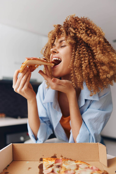 Jonge vrouw genieten van een heerlijk stuk pizza in de voorkant van een klassieke pizza doos met krullend haar - Foto, afbeelding