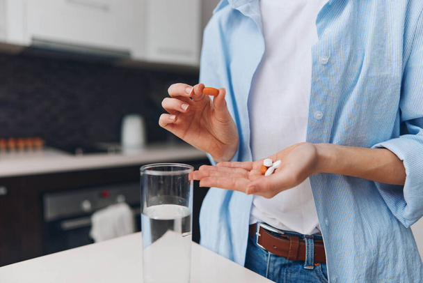 Женщина принимает лекарства со стаканом воды на кухне, стоящей перед прилавком - Фото, изображение