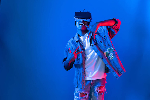 Usmívající se mladý Afroameričan dívající se skrz VR tanec v písni s hologramem hraní na růžové modré neonové stěně v metaverzním světě spojujícím digitální futuristickou technologii virtuální realitu. Přístroj. - Fotografie, Obrázek
