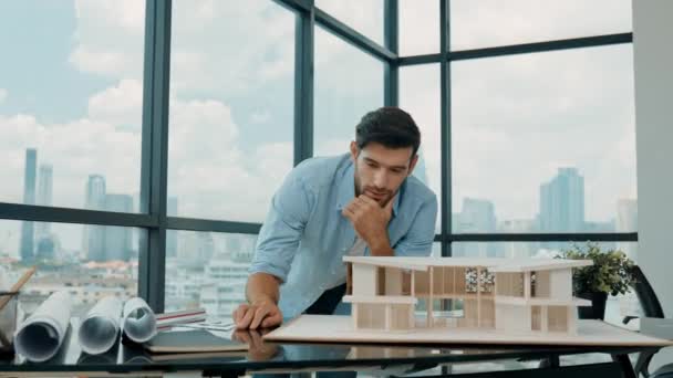 Hübsche kaukasische Ingenieure oder Projektmanager messen, inspizieren, schauen sich das Hausmodell an und schreiben auf den Bauplan, während sie in der Nähe eines Panoramafensters mit Stadt- und Wolkenkratzerblick stehen. Spurensuche - Filmmaterial, Video