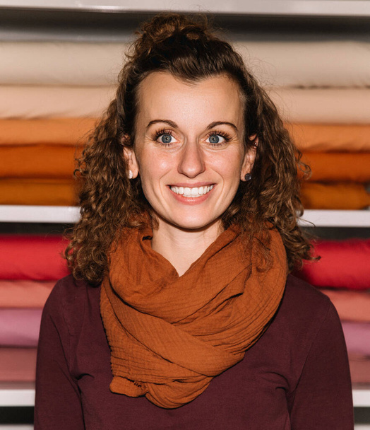 Portrét usměvavé módní návrhářky s kudrnatými vlasy stojící před úhledně uspořádanými barevnými látkovými rolkami ve svém ateliéru - Fotografie, Obrázek