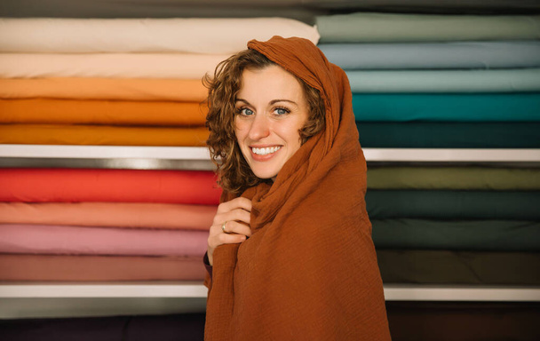  Une femme charmante souriante enveloppée dans une écharpe confortable dans une boutique de textile, concept de design - Photo, image