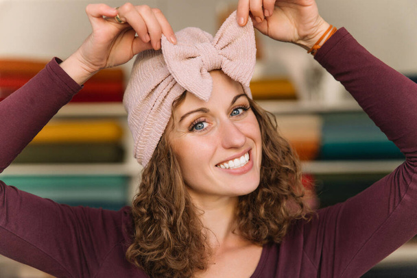 Radostná návrhářka si zkouší růžovou pletenou čelenku, její úsměv rozjasňuje kreativní pracovní prostor naplněný různými barevnými látkovými rolkami - Fotografie, Obrázek