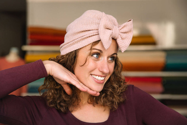 Joyful mode-ontwerper speels poseren met een handgemaakte roze strik hoofdband, haar hand als een nep-ondersteuning, in haar levendige stof beklede studio - Foto, afbeelding