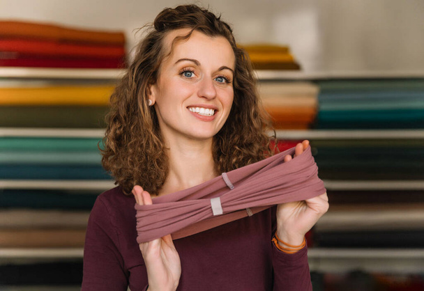 Créatrice de mode enthousiaste affichant fièrement un accessoire textile fraîchement fabriqué dans son atelier bien organisé, entourée de rouleaux de tissu colorés - Photo, image