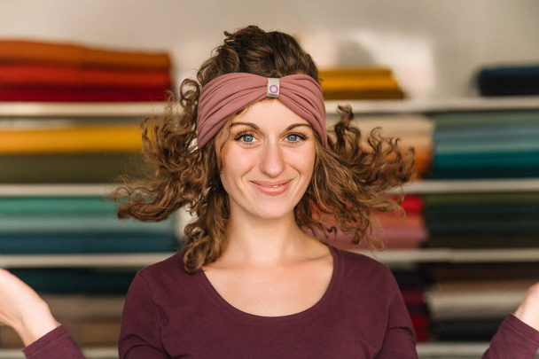 Μια χαρούμενη σχεδιάστρια μόδας με εκφραστικά μάτια και σγουρά μαλλιά ποζάρει παιχνιδιάρικα στο πολύχρωμο στούντιό της, επιδεικνύοντας περήφανα ένα chic headband - Φωτογραφία, εικόνα