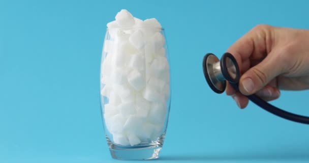 男の手は白い砂糖のキューブの完全なガラスに聴診器を置きます.医師は患者の健康への過度の砂糖消費の影響を調査する - 映像、動画