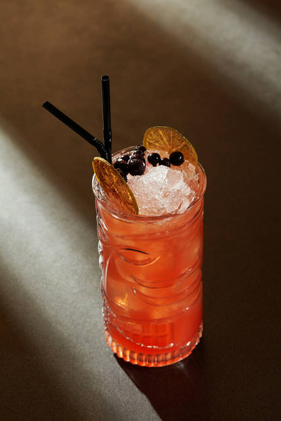 Szklanka orzeźwiającego tropikalnego koktajlu Zombie z owocowymi i pikantnymi nutami na bazie rumu z syropem cynamonowym i sokiem grejpfrutowym przyozdobionym jagodami i suszonymi plastrami wapna na ciemnym tle - Zdjęcie, obraz
