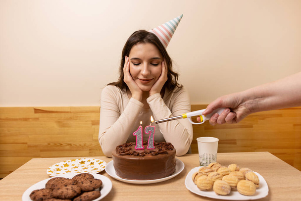 Дівчина сидить перед столом зі святковим тортом, в якому свічка освітлена у вигляді номера 17. Концепція святкування дня народження. - Фото, зображення