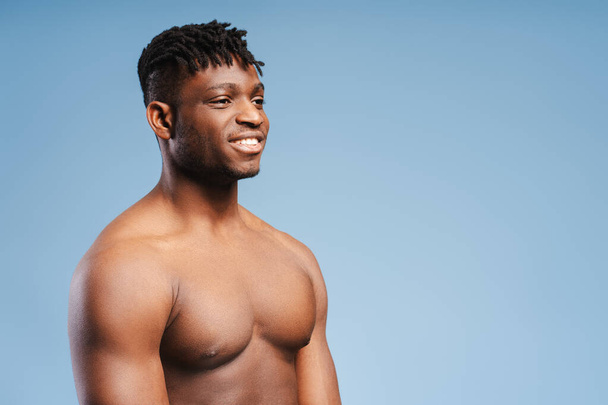 Smiling attractive African American man, athlete with bare, muscular torso looking away at copy space, standing isolated on blue background. Concept of advertising, shopping, healthy lifestyle - Photo, Image