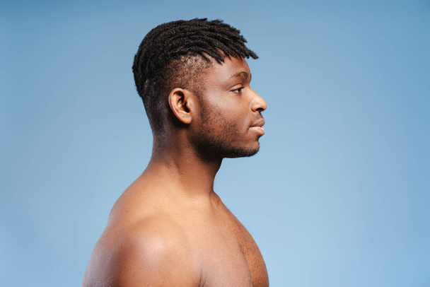 Side view, pensive african american man, fashion model with bare torso looking away at copy space, standing isolated on blue background. Concept of advertisement, shopping - Photo, Image