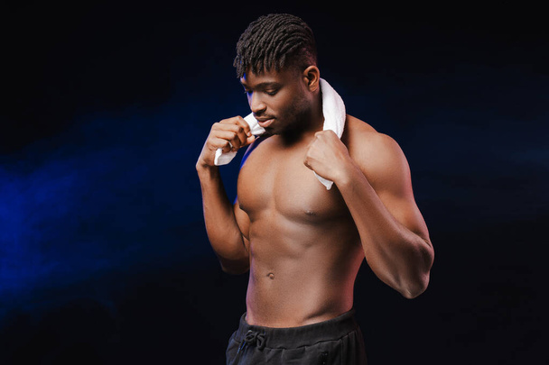 Handsome serious African American male athlete with bare pumped torso after workout holding towel standing isolated on black background. Concept of healthy lifestyle, exercise, sport - Photo, Image