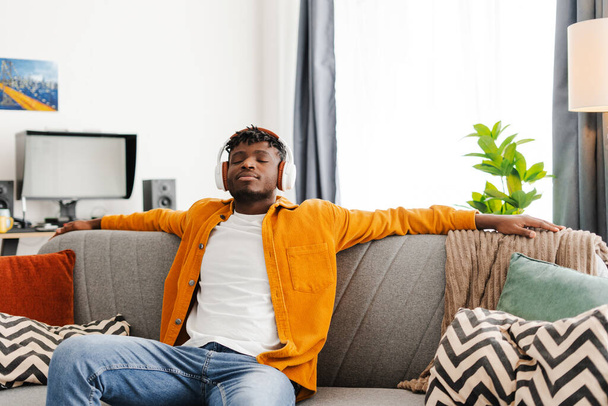 Alegre hombre afroamericano escuchando música en los auriculares, disfrutando de sentarse en casa acogedora. Concepto de tecnología, hobby - Foto, imagen