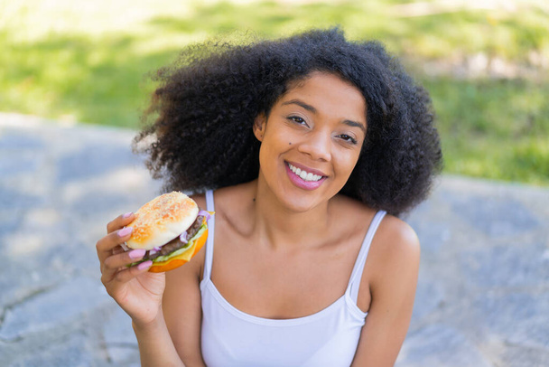 Jonge Afro-Amerikaanse vrouw houdt een hamburger in de buitenlucht glimlachen veel - Foto, afbeelding