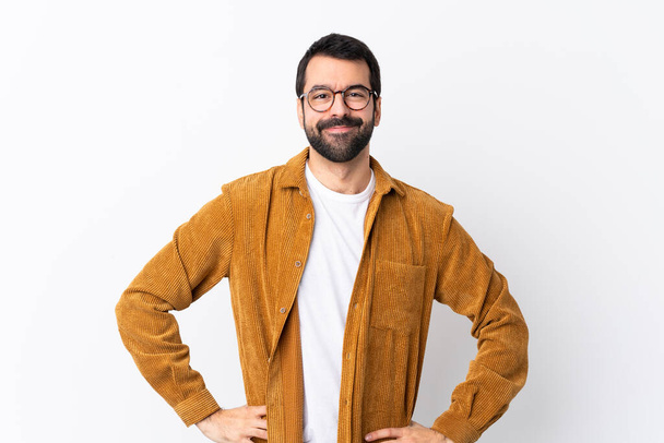 Kavkazský pohledný muž s vousy v manšestrové bundě přes izolované bílé pozadí pózuje s pažemi v bok a usmívá - Fotografie, Obrázek